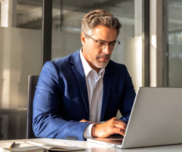 A man in a blue suit sits at his desk searching on his laptop
